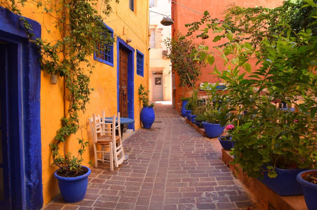 A narrow cobbled lane in Chania between saffron-yellow walls, cobalt doors and blue pots of plants