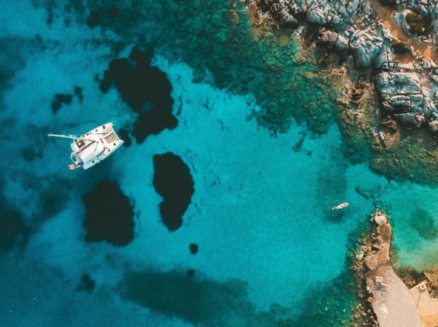 A white catamaran anchored in a turquoise cove, seen from above, beside rocky shore