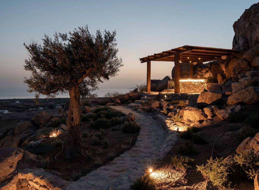 An olive tree and a lit stone path leading to a pergola at dusk above the Aegean