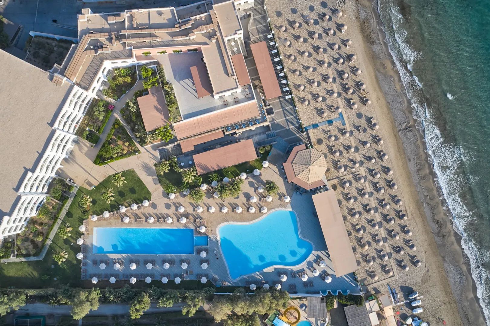 An aerial view of the resort's two pools beside a long beach lined with umbrellas and the turquoise sea