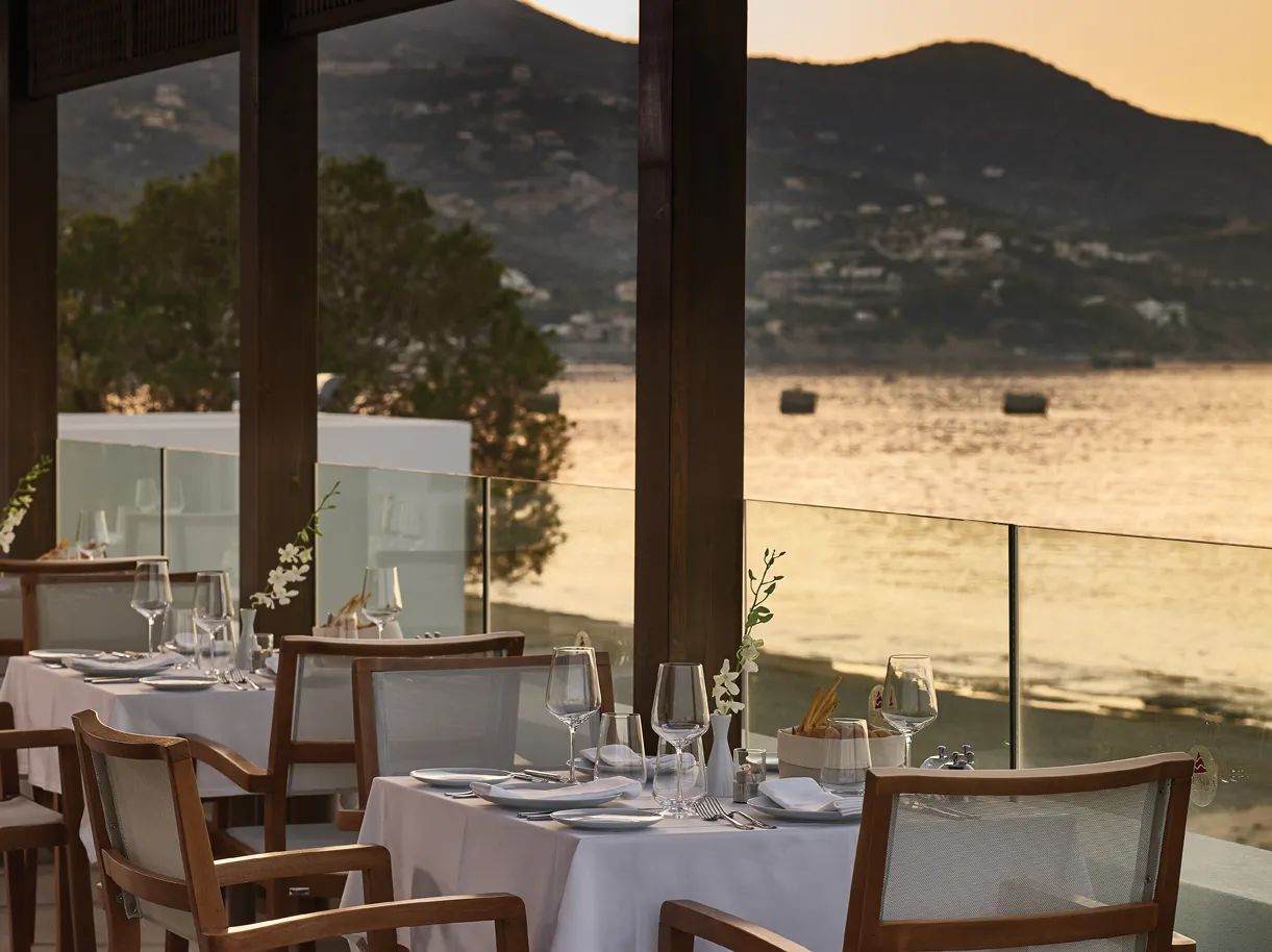 Tables set with white linen on a glass-railed terrace overlooking the bay at sunset