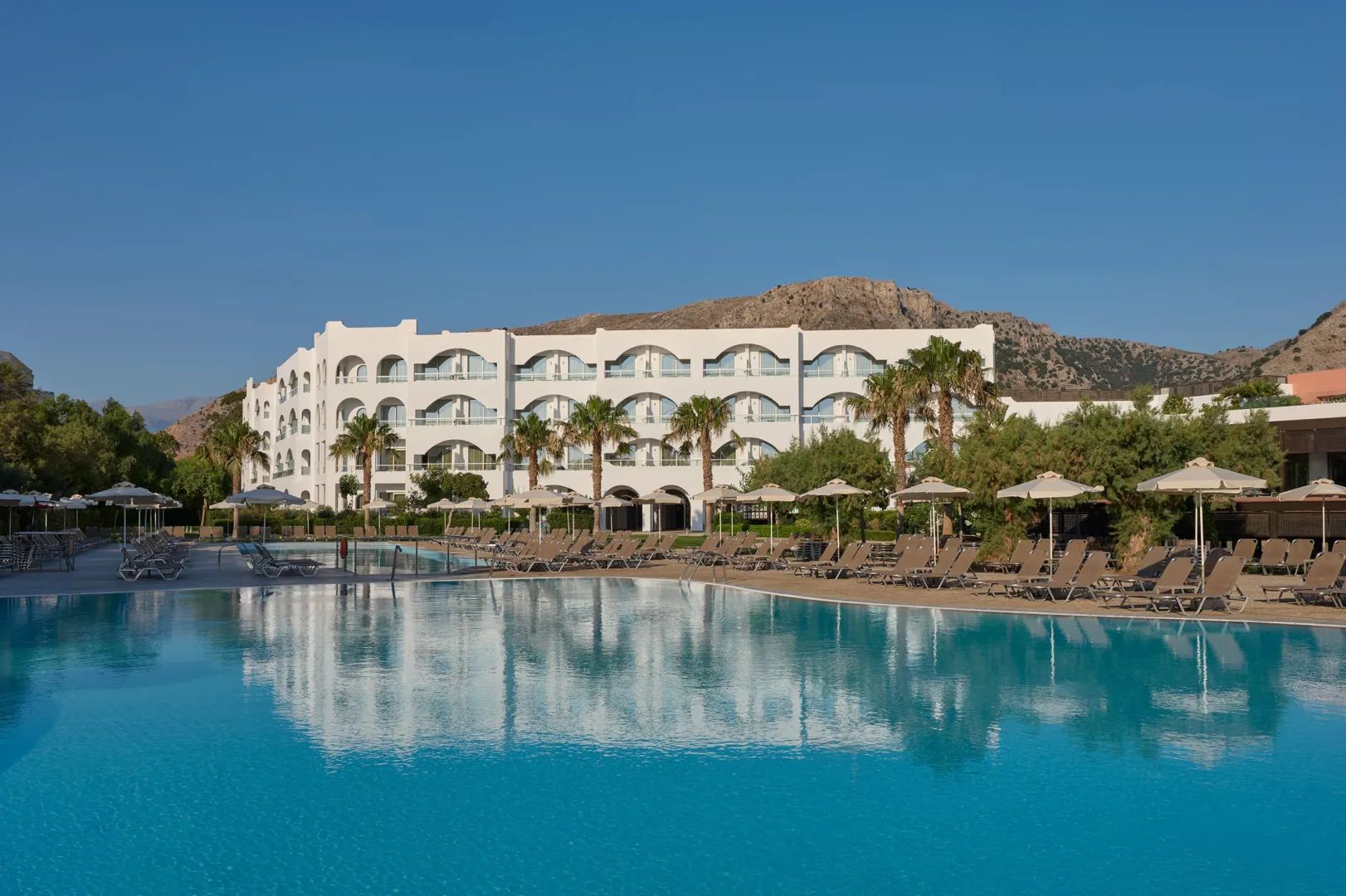 A white arched resort building reflected in a still turquoise pool, mountains rising behind