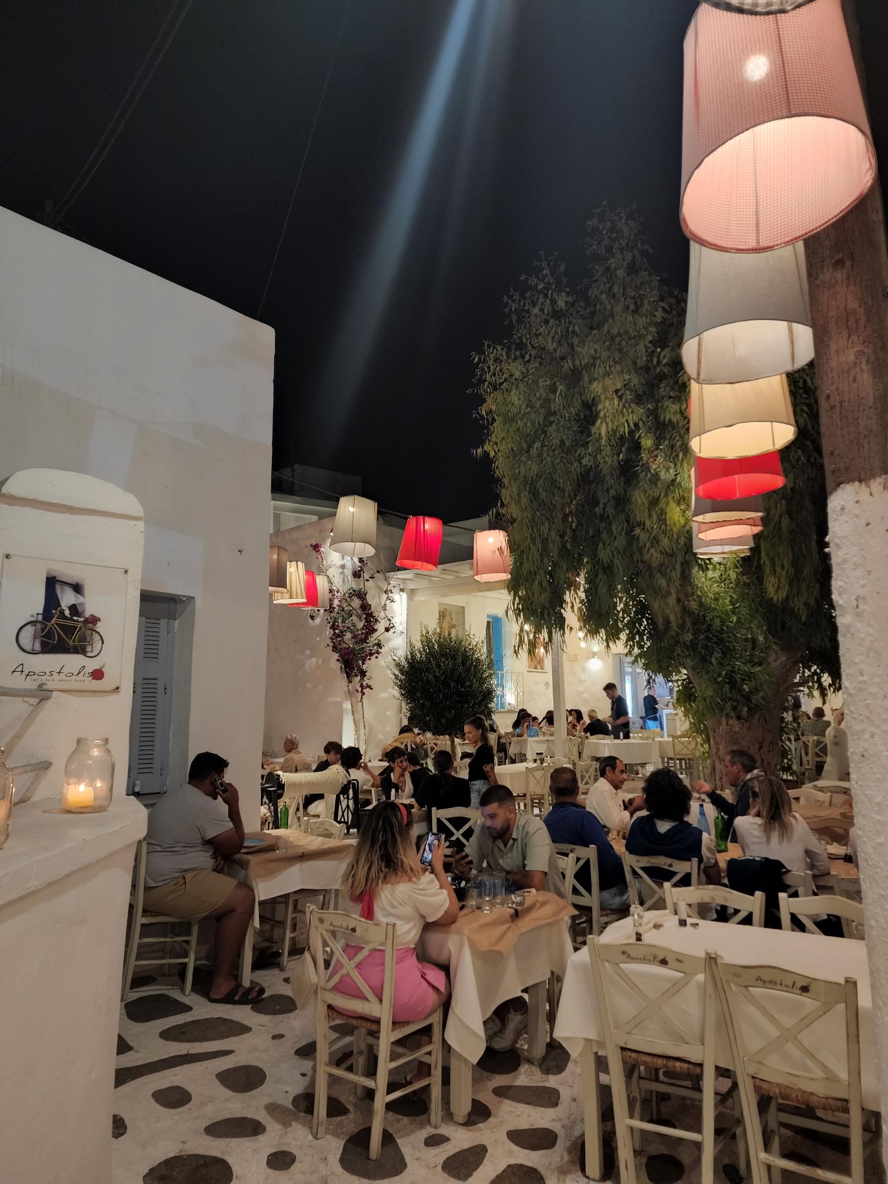 A lantern-lit taverna square at night, tables under a tree hung with pink and red lamp shades