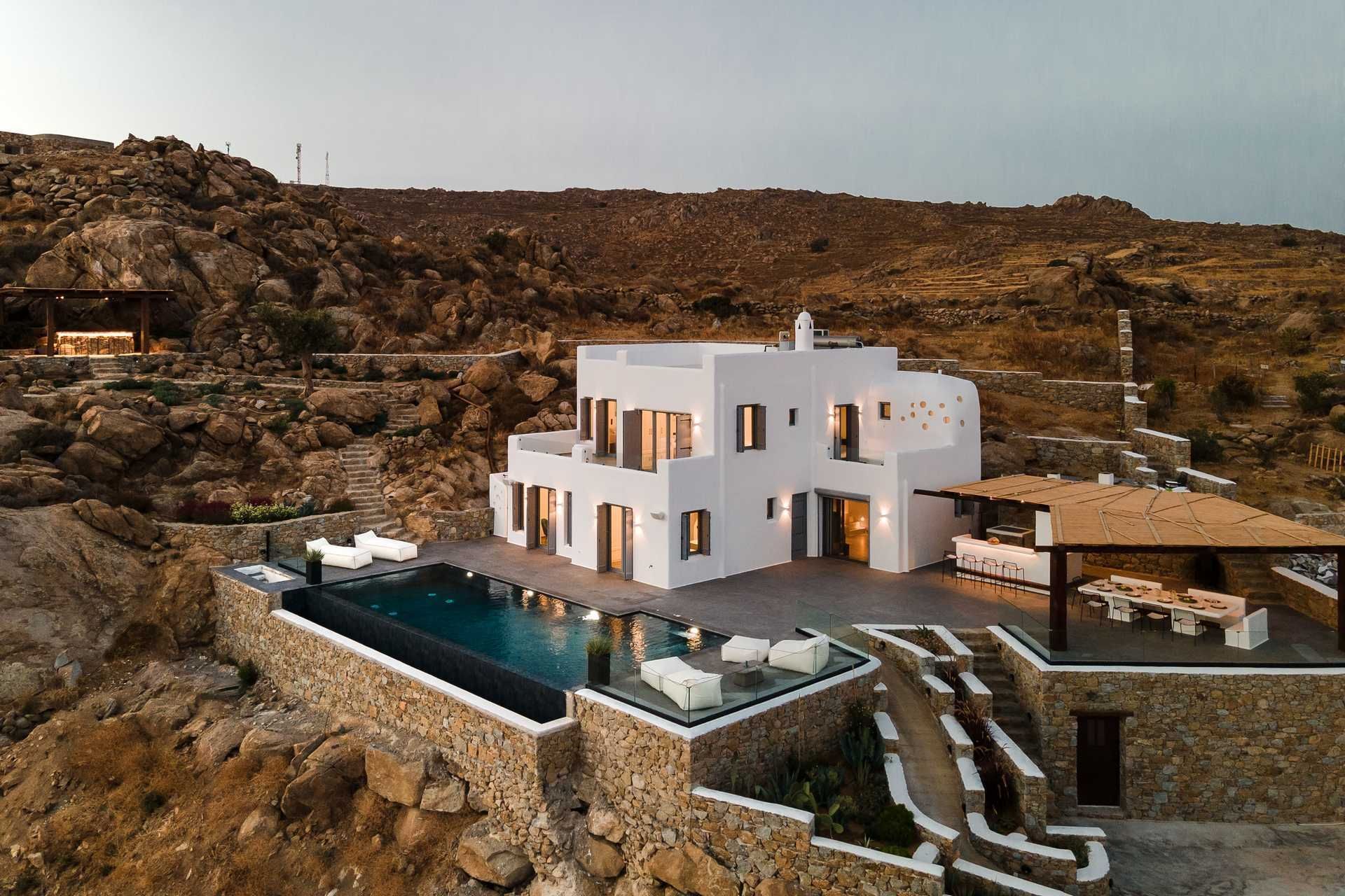 A whitewashed hilltop villa with an infinity pool and reed-roofed terrace, glowing at dusk over the rocky hills of Naxos