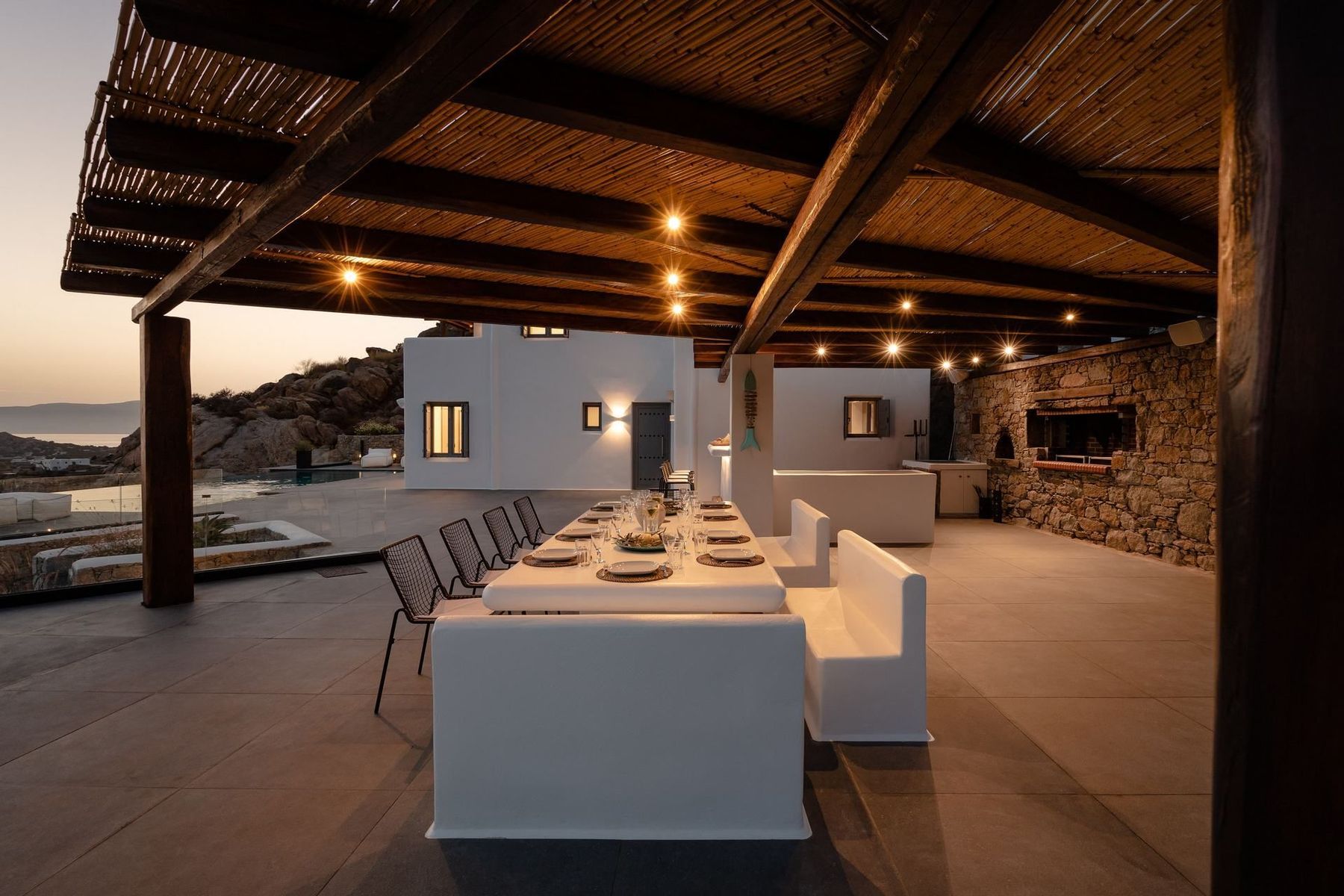 The pergola at dusk with string lights glowing over the dining table and the stone outdoor kitchen